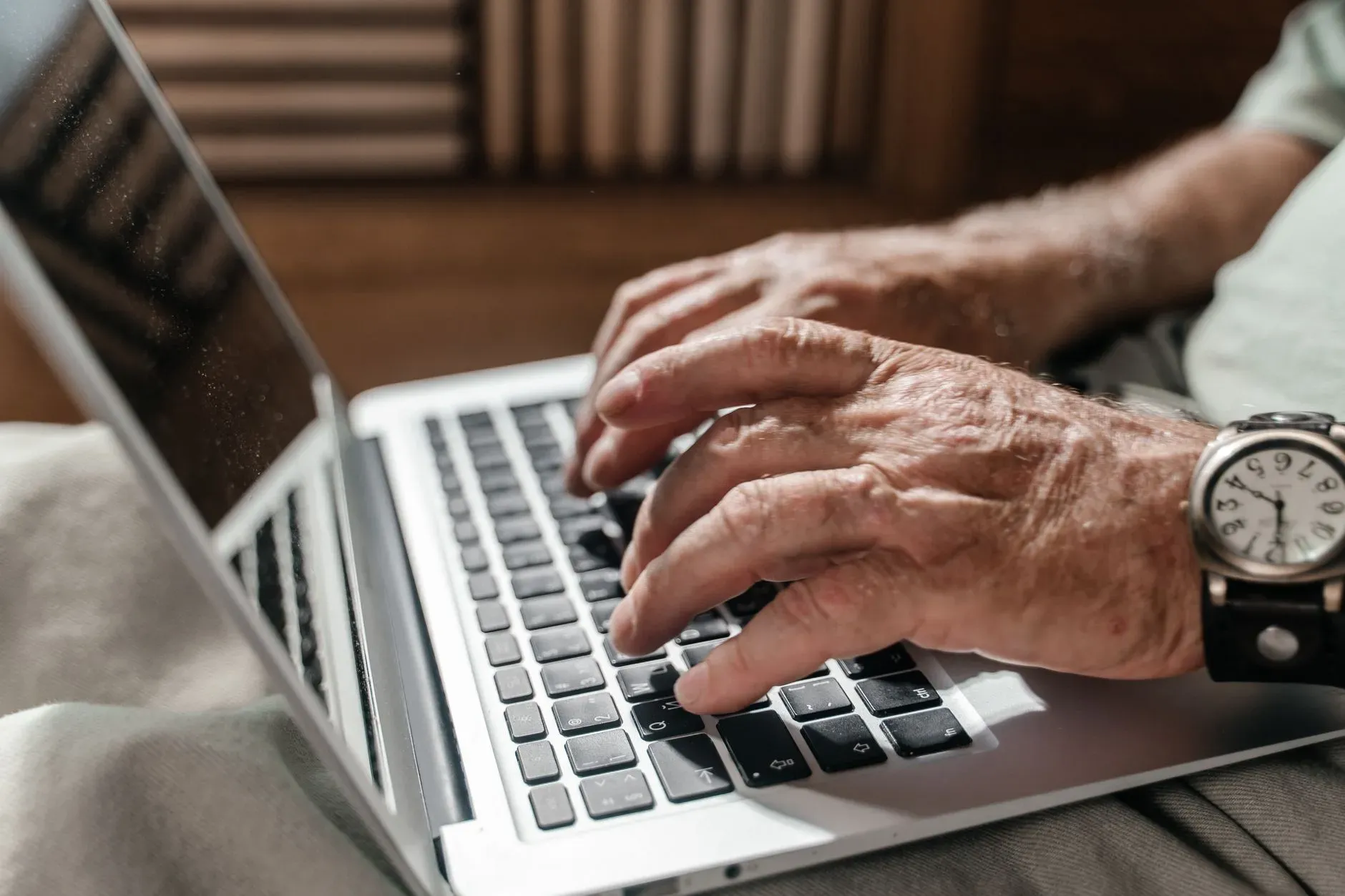 Primo piano delle mani di una persona anziana che digita su un computer portatile e indossa un orologio da polso, in un ufficio a casa.